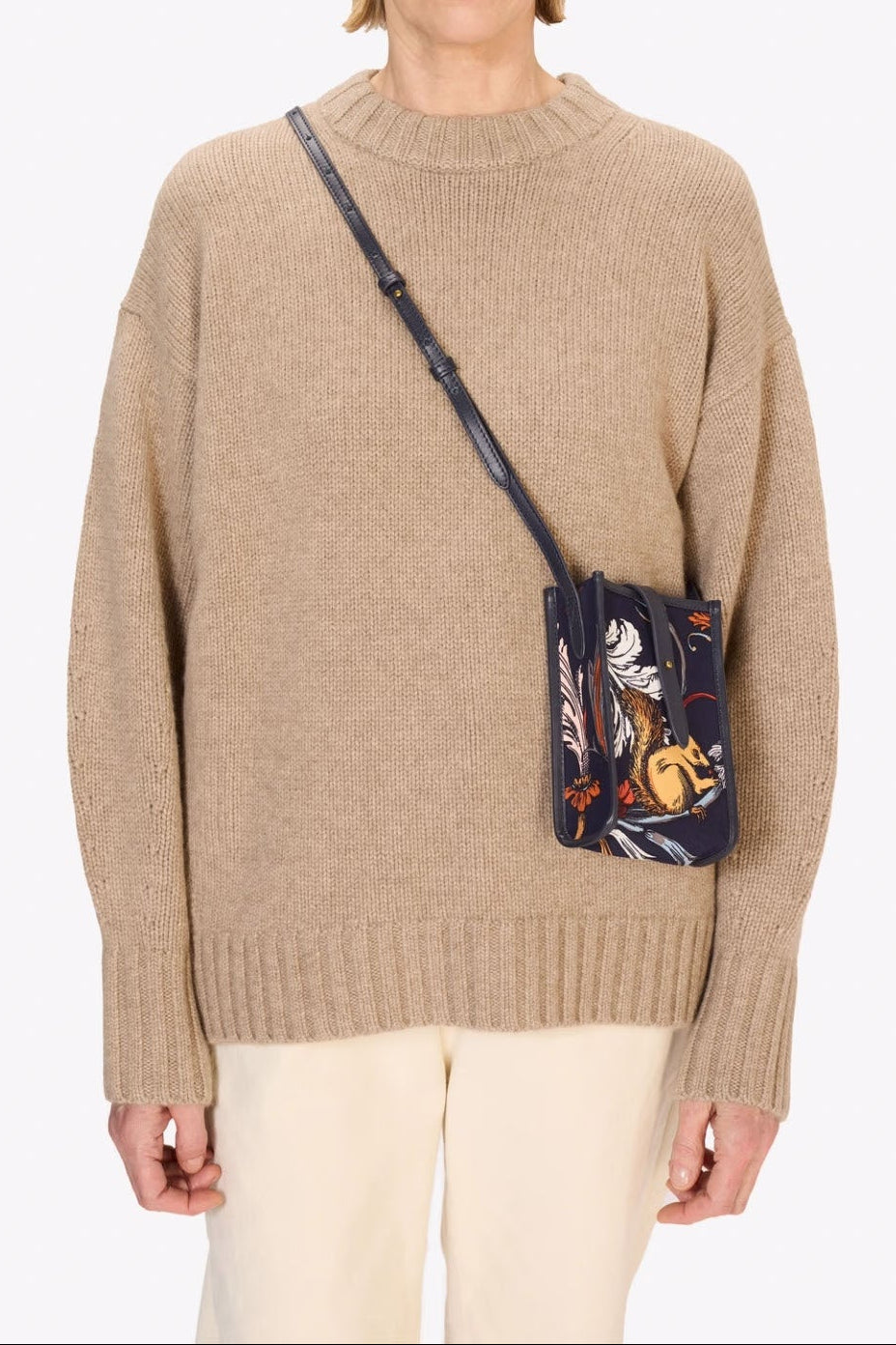 A woman wearing a beige knit sweater and cream pants, carrying a small dark crossbody bag with a colorful floral print and a black strap, against a plain light gray background.
