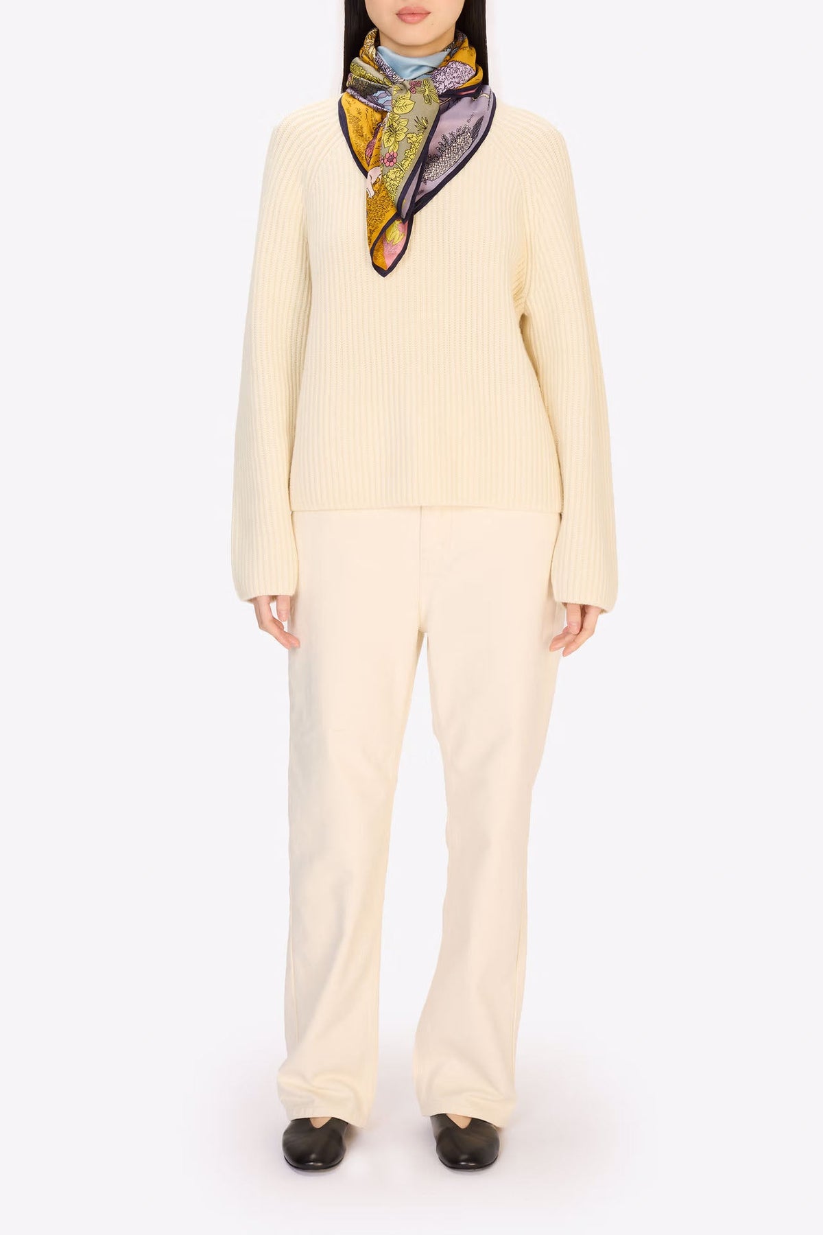 A model stands against a plain white studio background wearing a cream-colored knit sweater and matching wide-leg trousers, accessorized with a patterned scarf in yellow, blue, and purple.