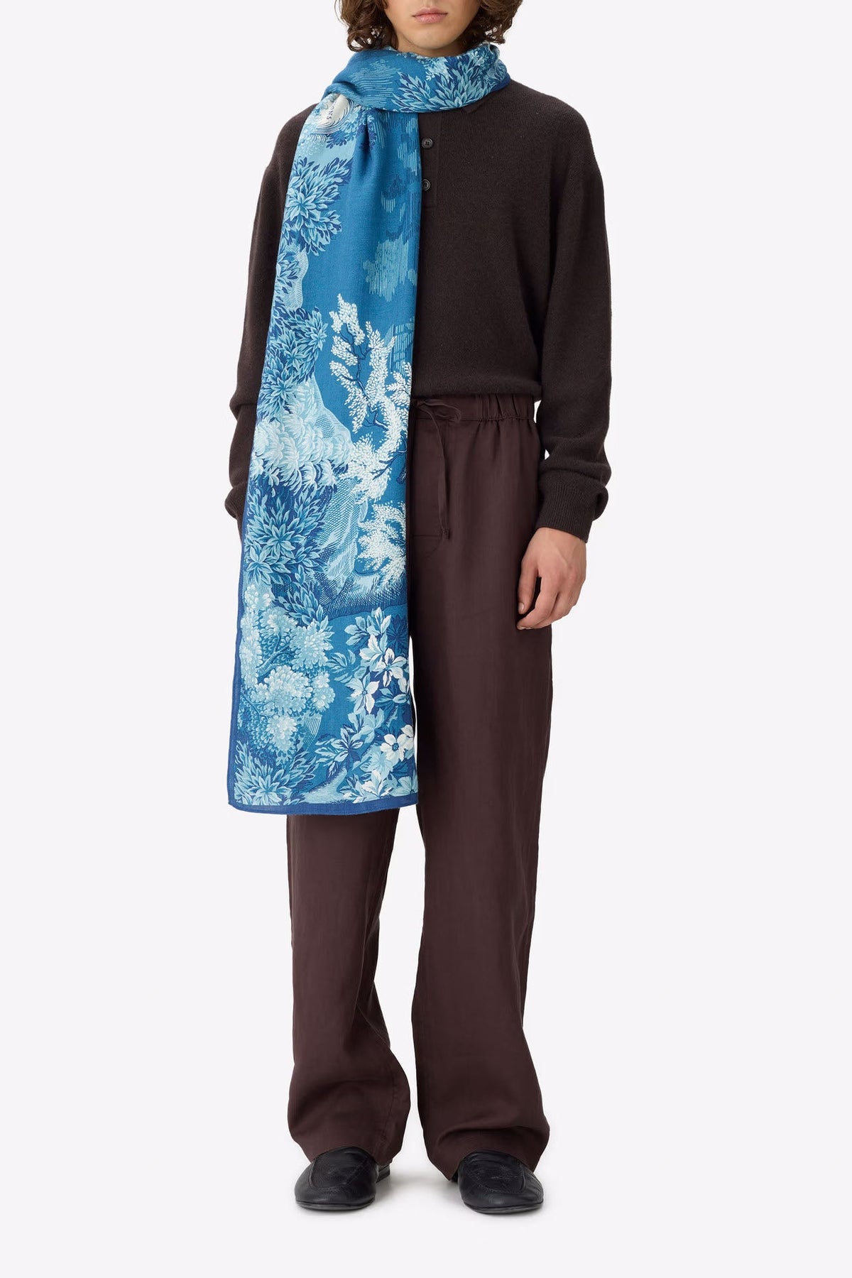 A model stands against a light gray studio background wearing a dark brown knit zip-up sweater, matching wide-leg pants, and a long blue patterned scarf draped over the shoulders.
