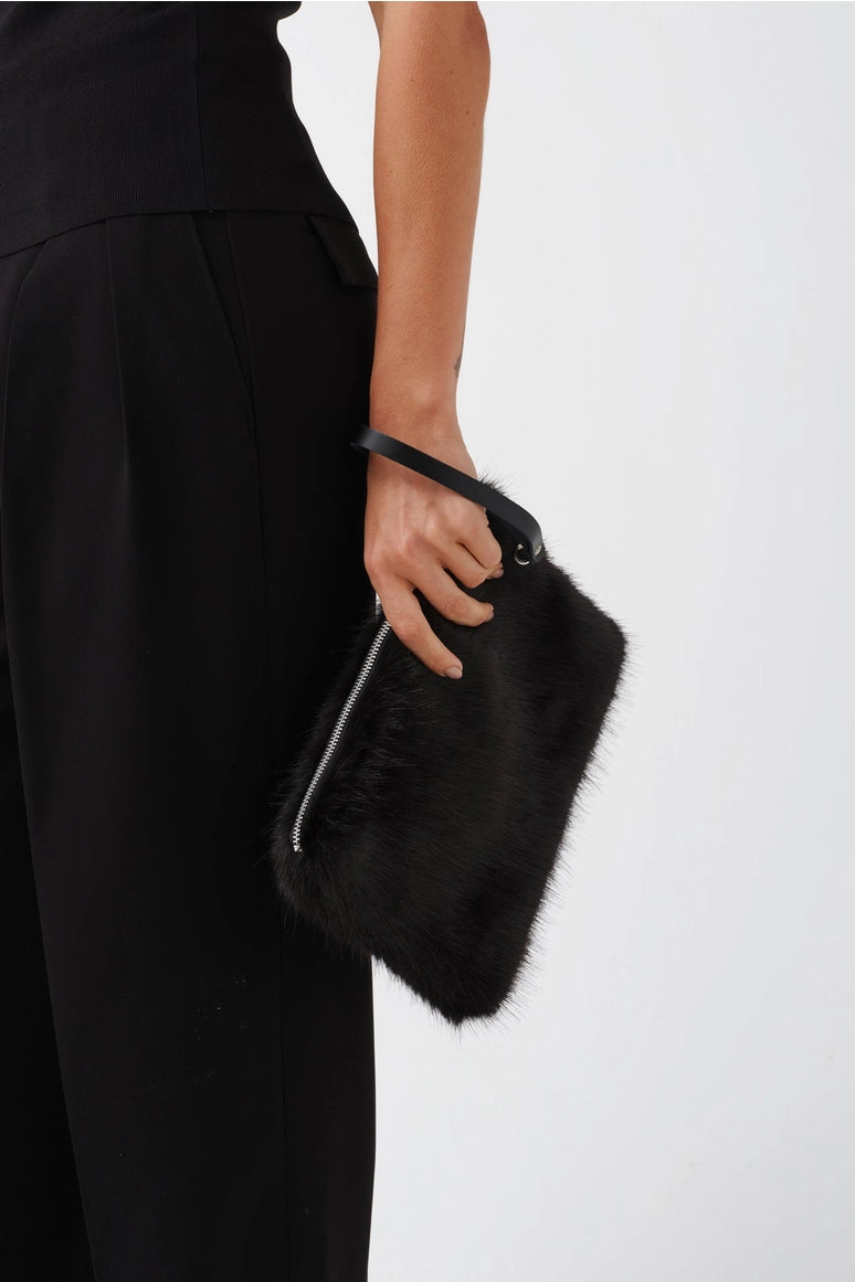 A person in a black outfit holds a black furry clutch bag with a black strap and silver zipper against a plain light gray background.