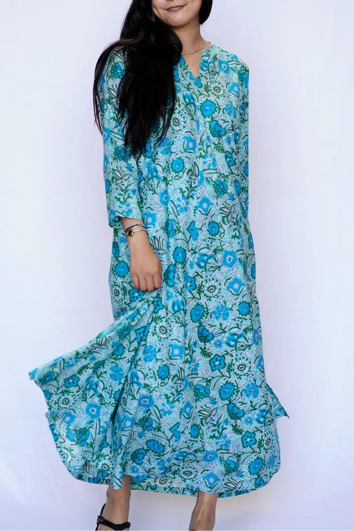 A woman modeling a long-sleeved, V-neck maxi dress with a blue and green floral block print on a light blue base, set against a plain white studio background.