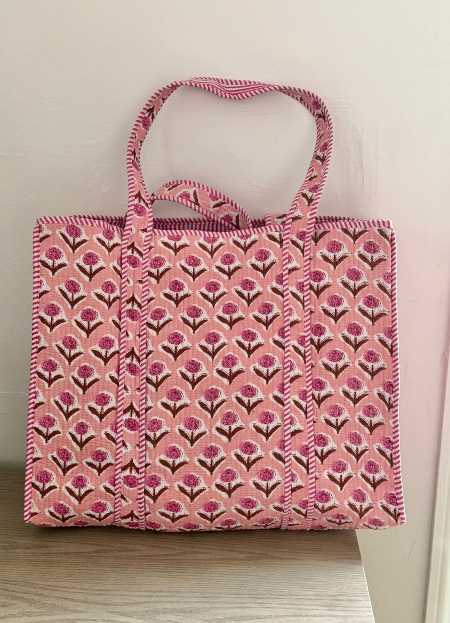A pink tote bag with a repeating floral block print pattern in pink, purple, and brown, featuring dual top handles, resting on a light wood surface against a plain off-white wall.