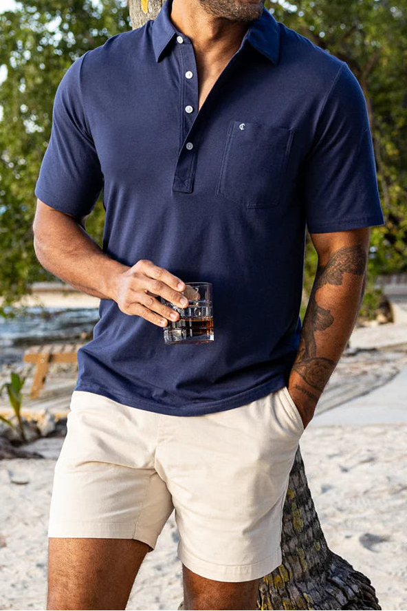 Man in navy polo shirt and beige shorts standing by a beach with trees and ocean in the background