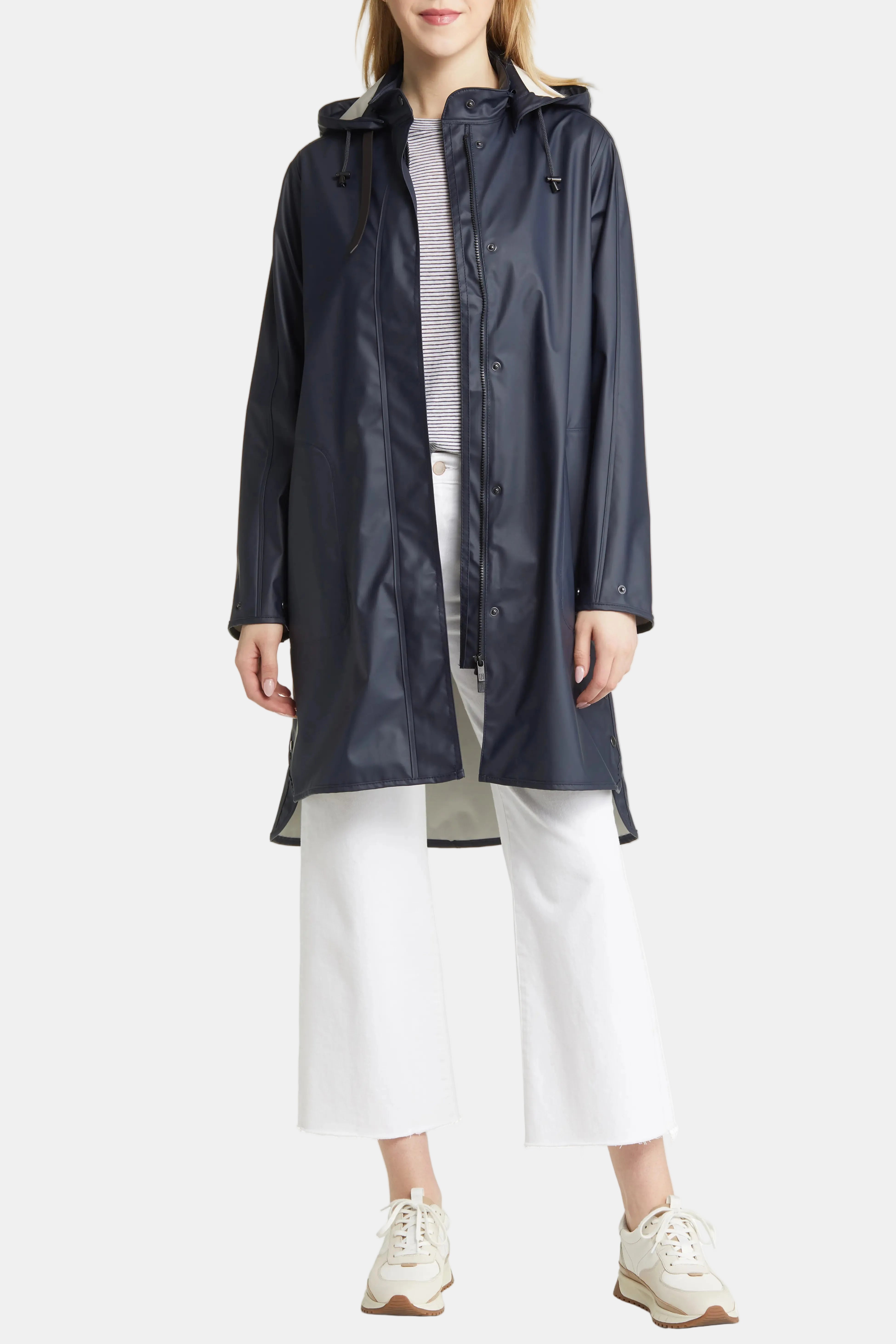 A model wearing a dark navy hooded raincoat over a gray striped shirt, white cropped pants, and white sneakers, photographed against a plain white studio background.