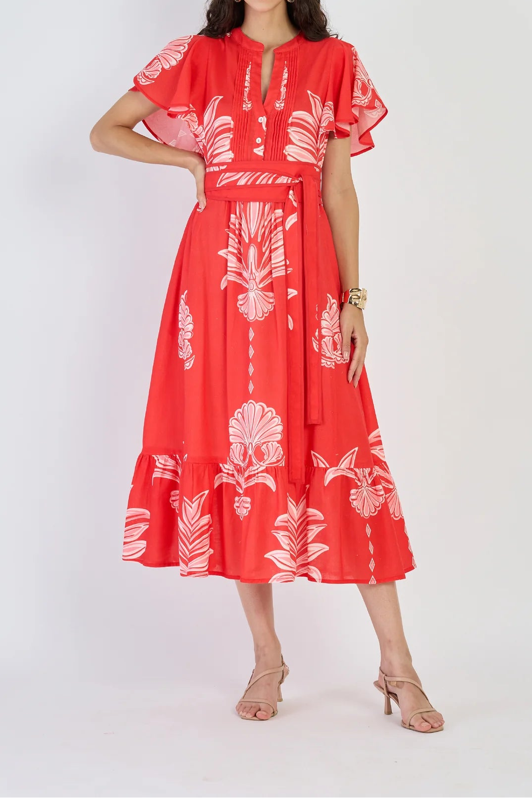 A woman modeling a red midi dress with a white tropical floral print, featuring short flutter sleeves, a tie-neck detail, and a matching waist tie, set against a plain light gray background.