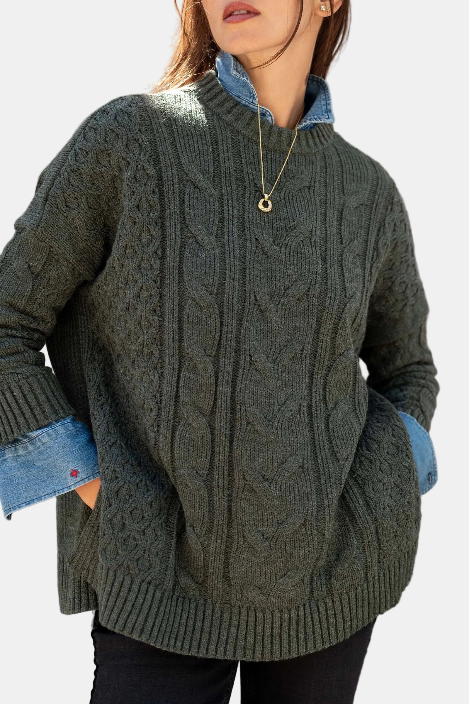 A woman wearing an oversized dark olive green cable-knit sweater over a light blue denim collared shirt, standing outdoors against a stone wall.
