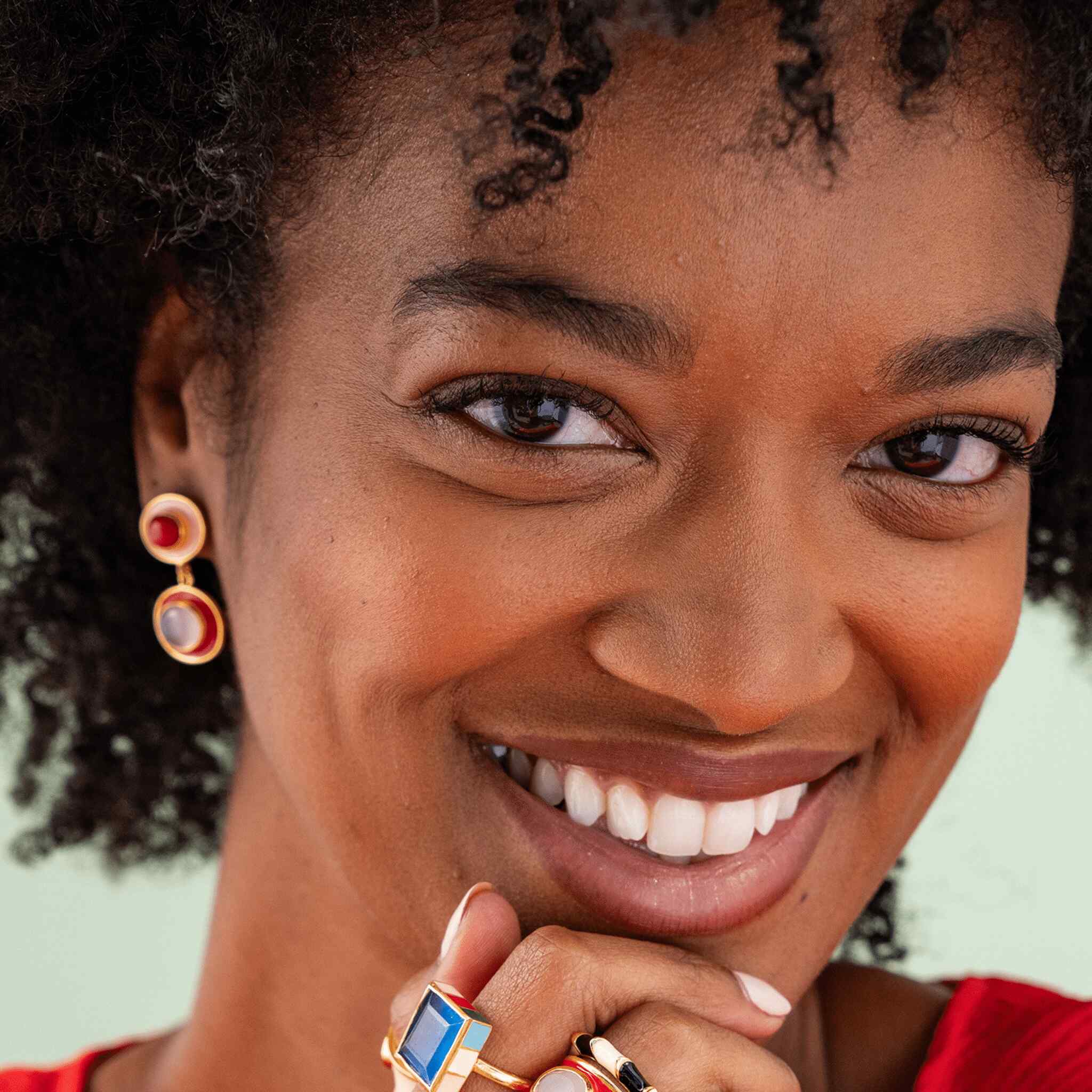 Close-up portrait of a smiling woman with dark curly hair wearing gold-toned drop earrings with red and white circular details, and multiple rings on her fingers, against a pale mint background.