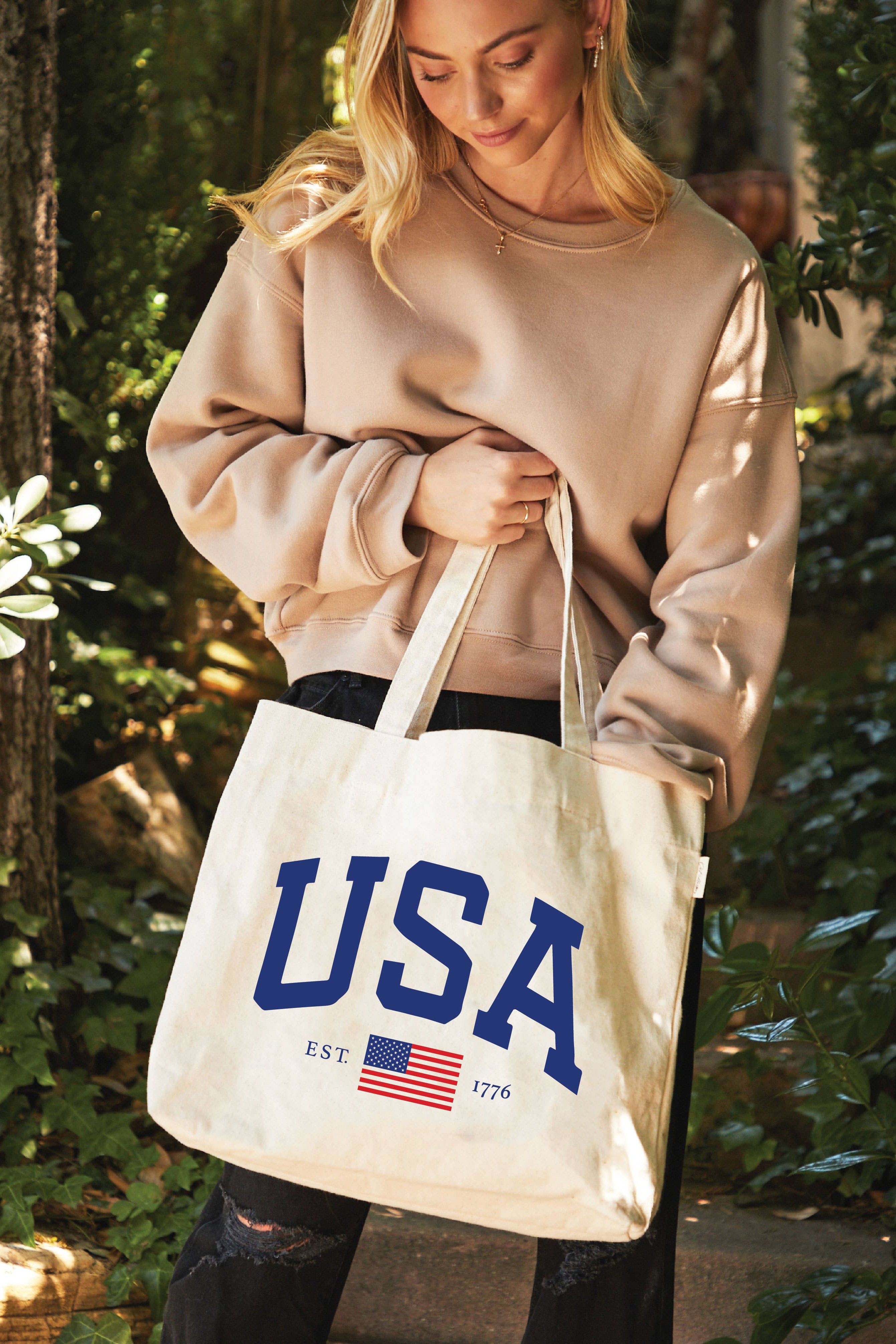 Woman holding a tote bag with 'USA' and an American flag design outdoors.
