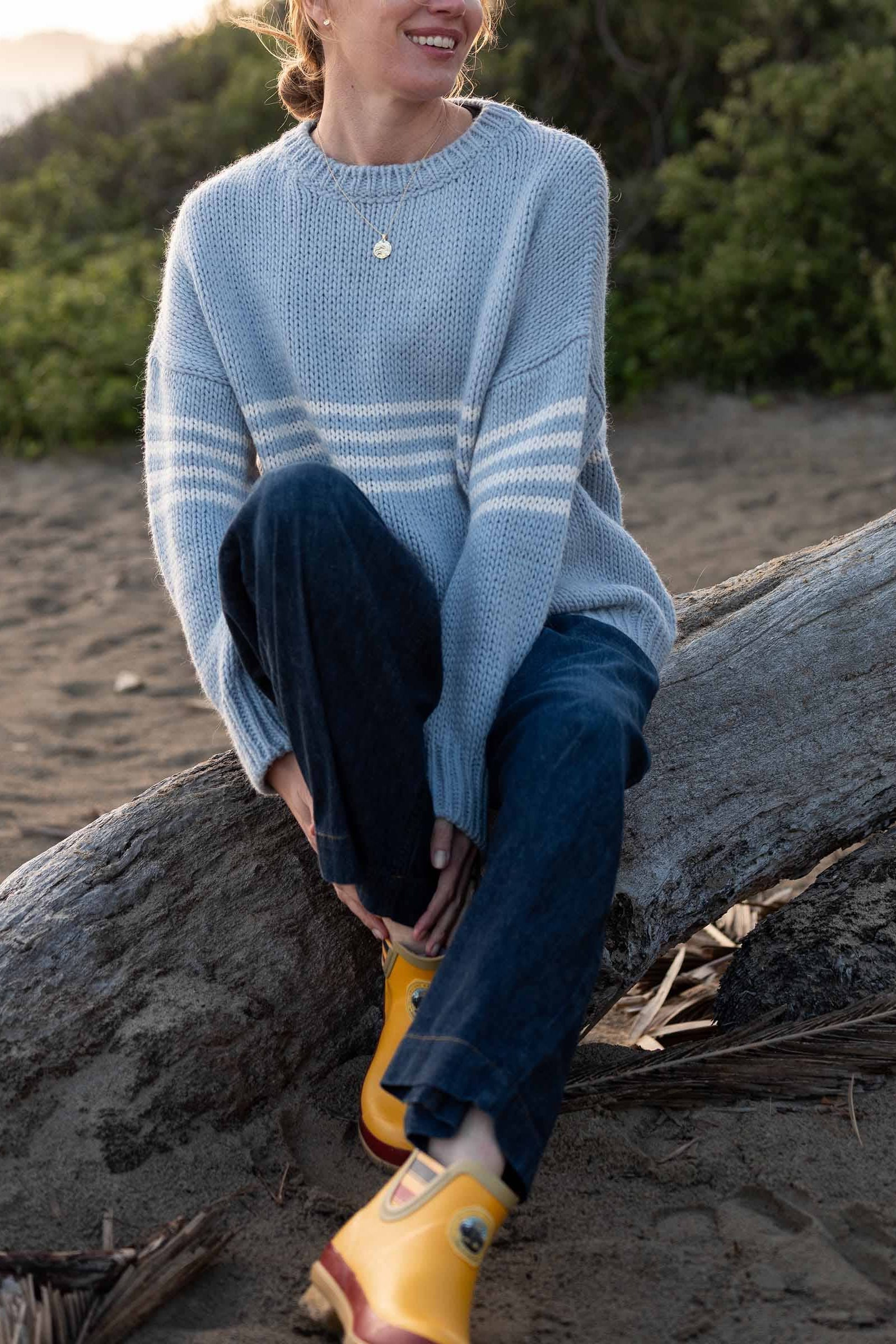 Person sitting on a log on a beach wearing a blue sweater and yellow boots.