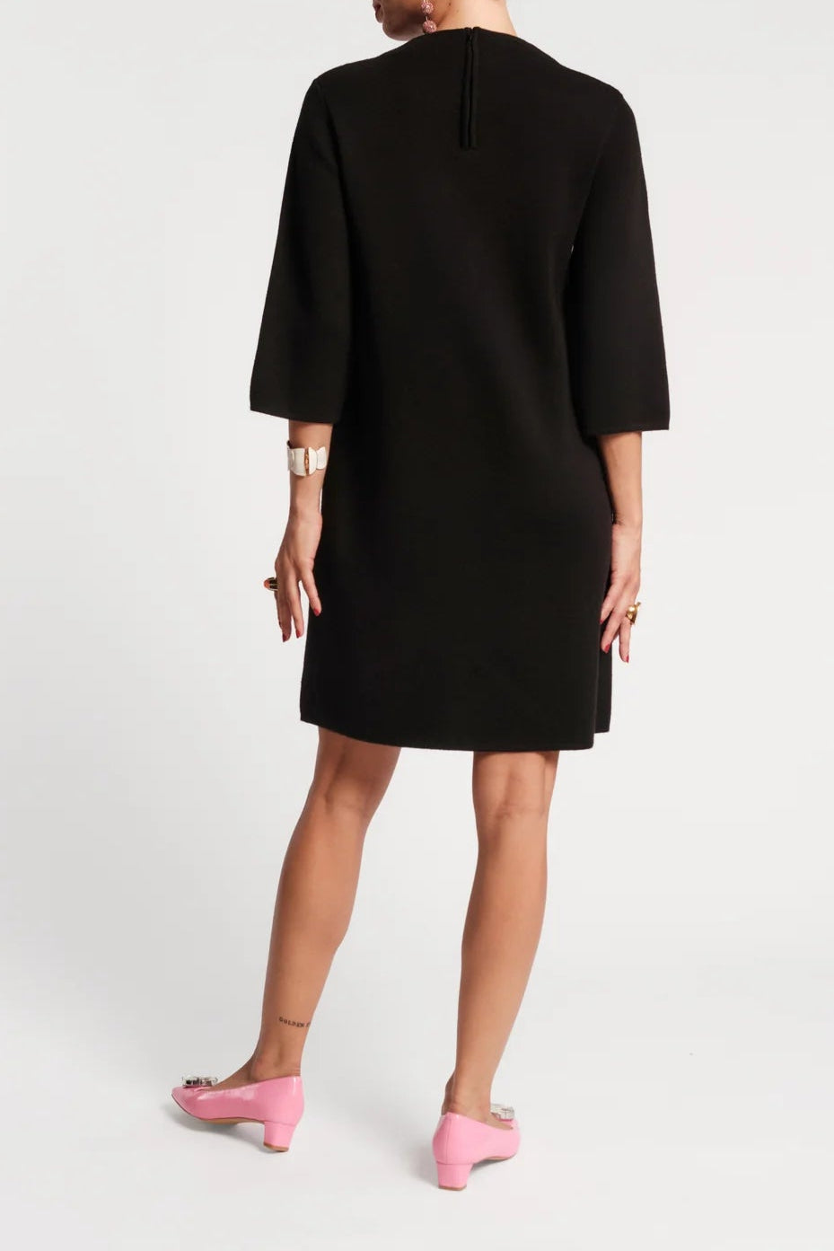 A woman shown from the back wearing a black three-quarter sleeve shift dress and pink pointed flats against a light gray studio background.