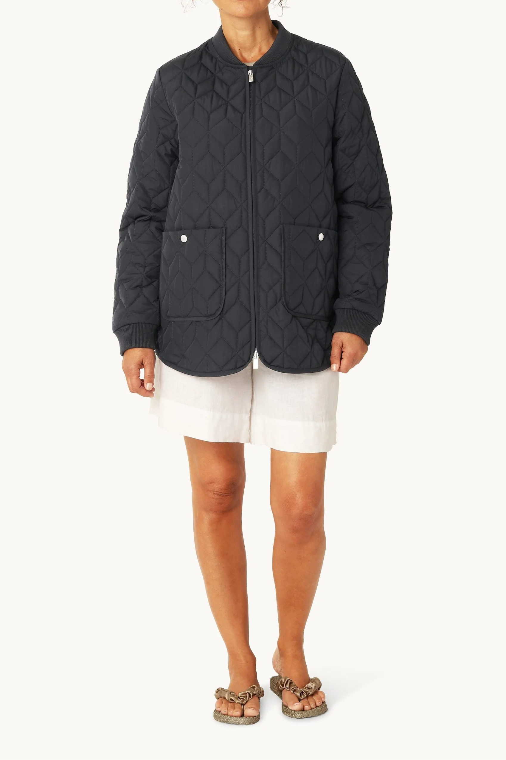 Woman wearing a dark quilted jacket and white shorts on a light gray background