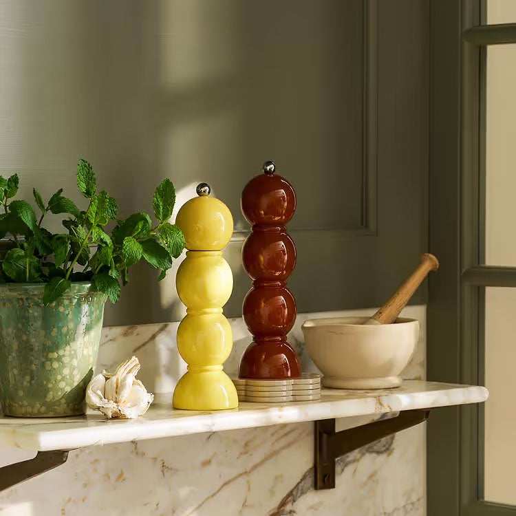 Two decorative stacked glass spice grinders, one yellow and one amber-brown, displayed on a white marble shelf next to a potted green plant and a cream mortar and pestle.
