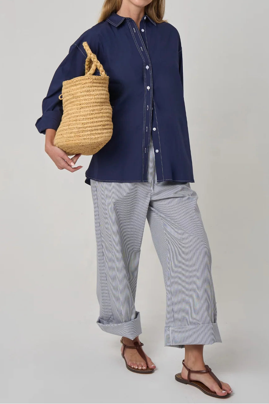 A woman modeling a navy blue long-sleeve button-up shirt, light blue and white wavy striped wide-leg pants, brown leather sandals, and holding a natural straw tote bag against a light gray studio background.