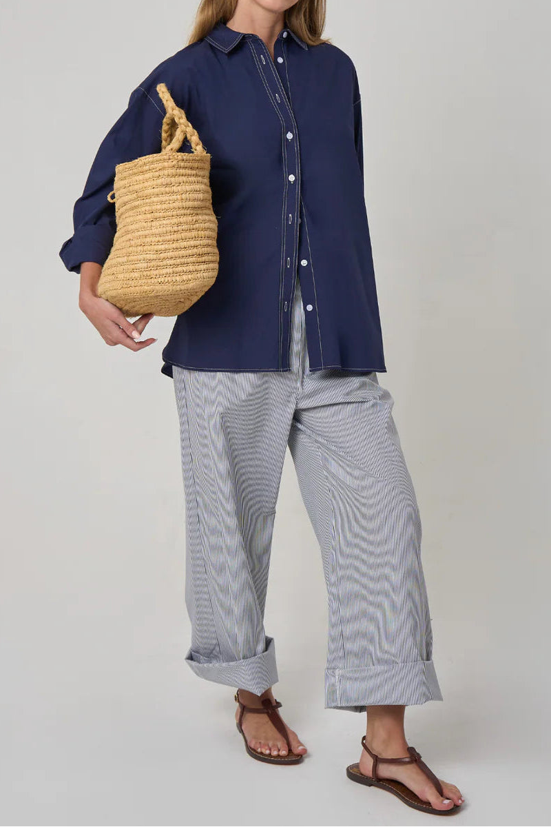 Woman holding a woven bag, wearing a navy shirt and light gray pants, on a plain background