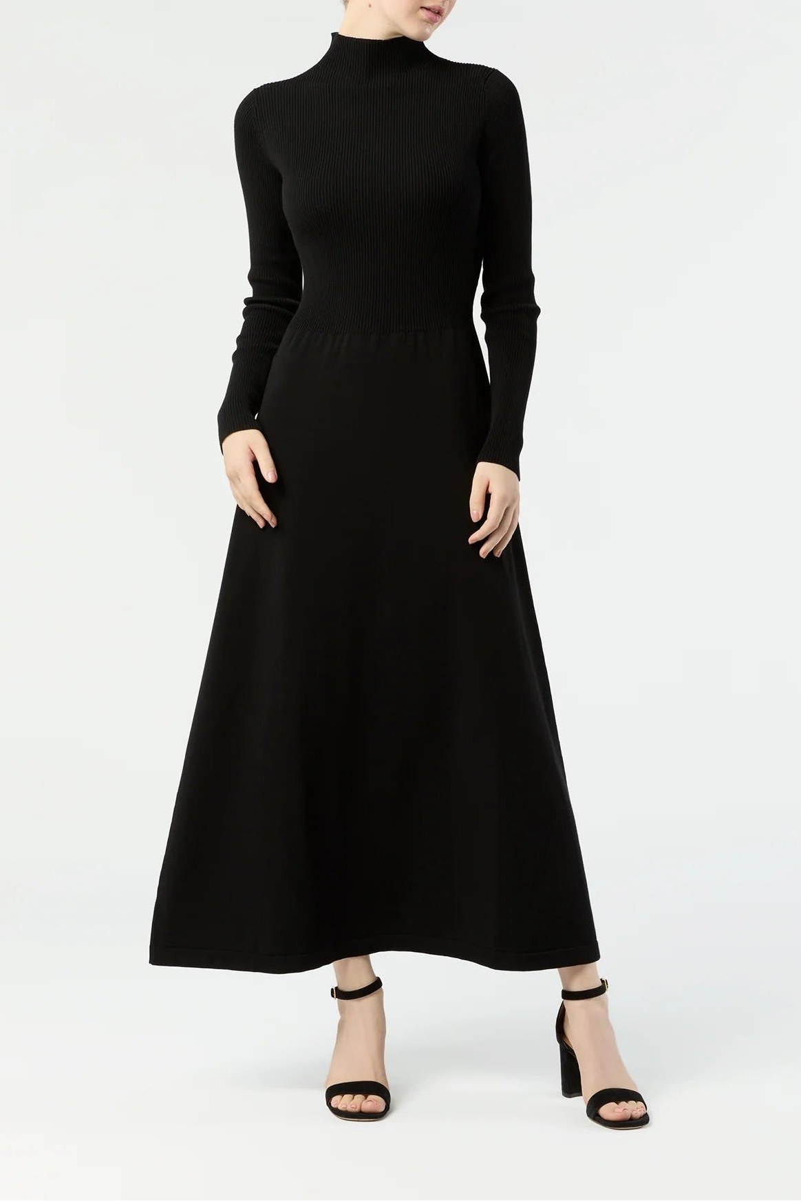 A woman modeling a black long-sleeve mock neck midi dress with a flared skirt, paired with black heeled sandals, against a plain white studio background.
