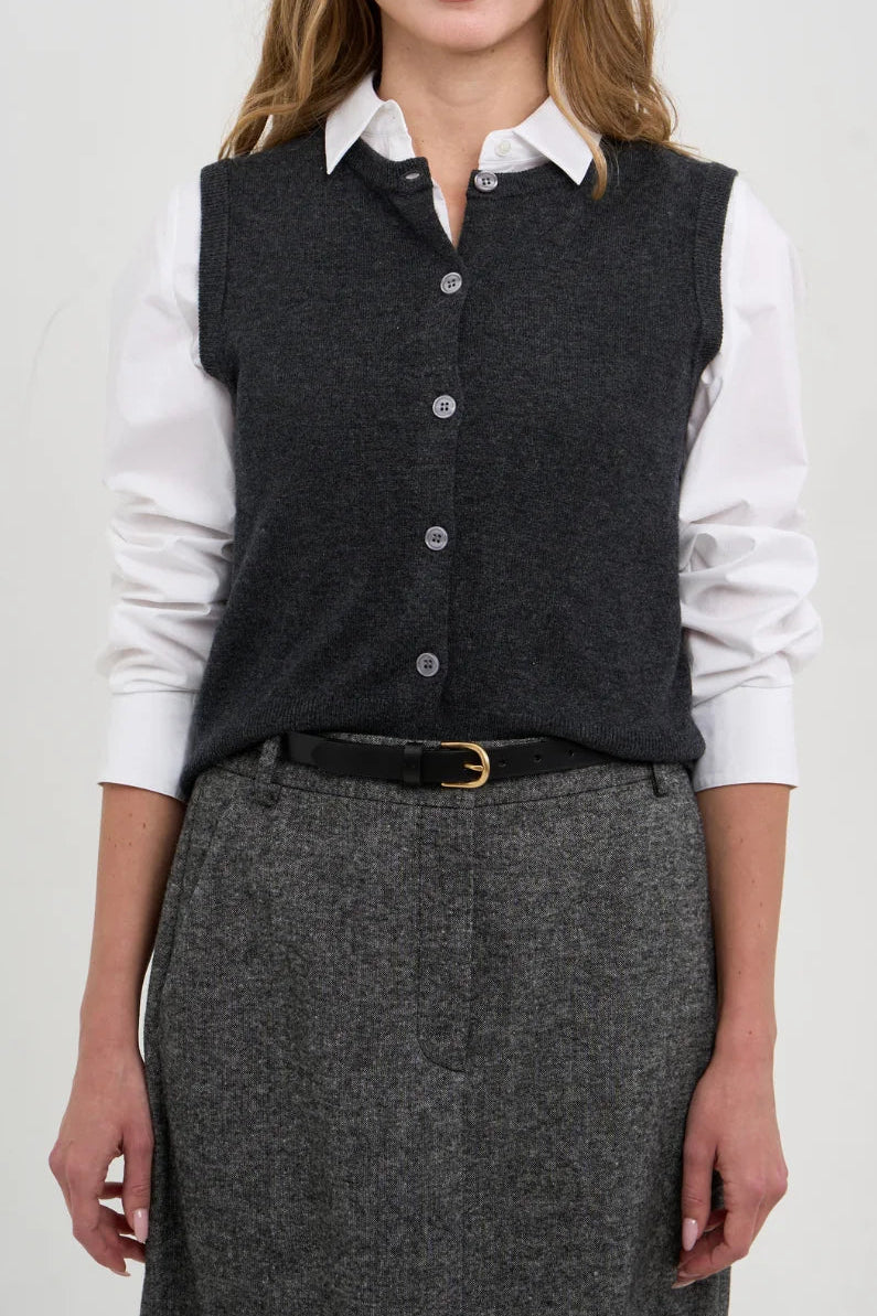 A woman modeling a dark gray button-front knit vest over a white collared shirt, paired with a gray skirt and black belt, against a plain light gray studio background.