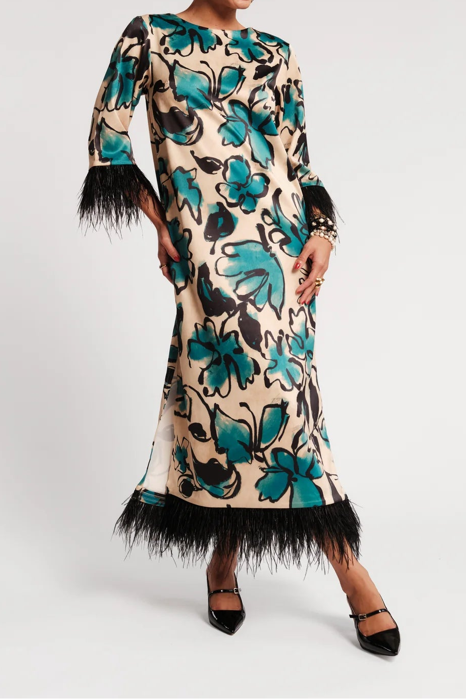 A woman models a beige midi dress with a teal and black floral print, featuring black feather trim at the hem and three-quarter sleeves with feather accents, set against a plain light gray background.