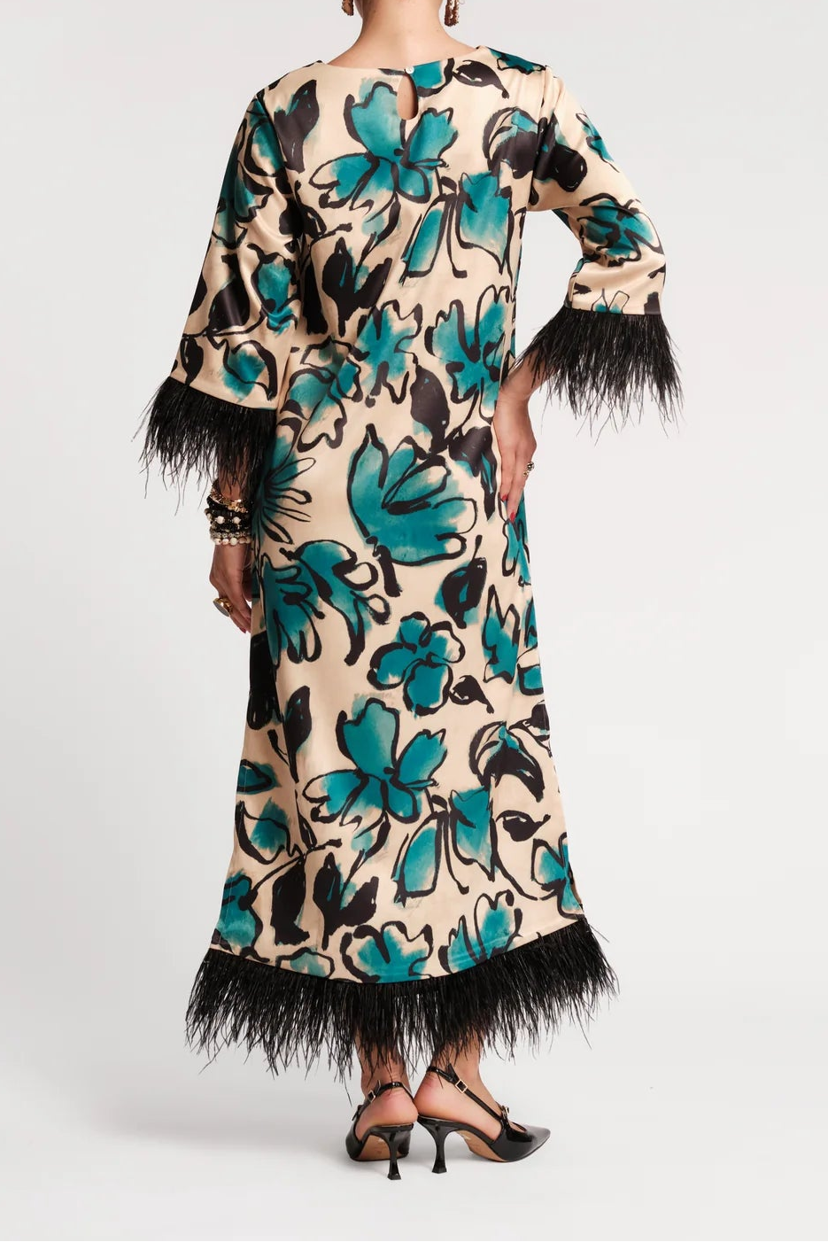 A woman viewed from the back wearing a long beige dress with a bold teal and black floral print, black feather trim at the sleeves and hem, and black strappy heels against a light gray studio background.