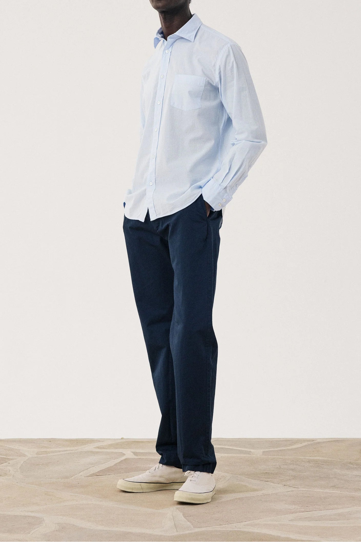 Man wearing a light blue shirt and dark pants standing against a white background