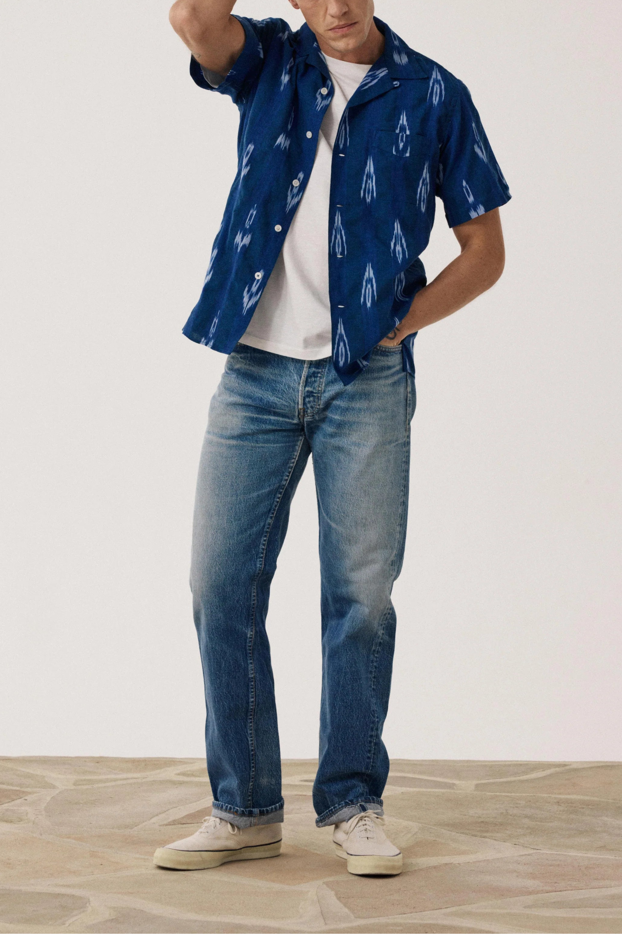 Man wearing a blue patterned shirt and jeans standing against a plain background