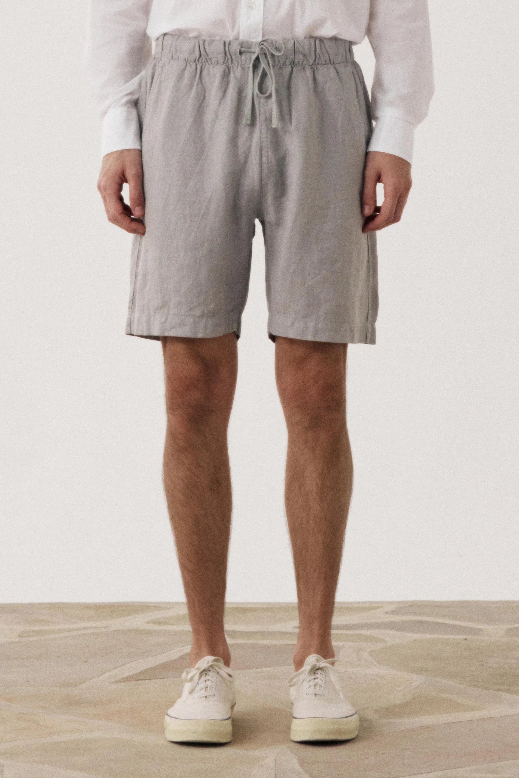A person standing against a light neutral wall wearing light gray drawstring shorts, a white long-sleeve shirt, and white sneakers.