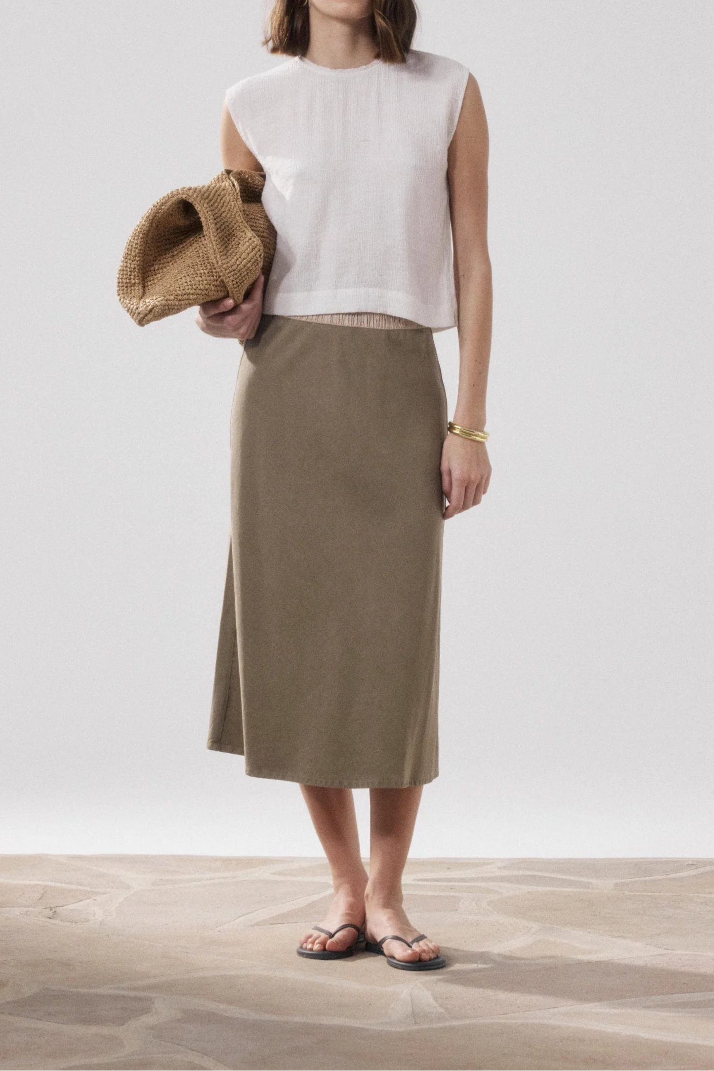 Woman wearing a white sleeveless top and beige skirt holding a woven bag on a plain background