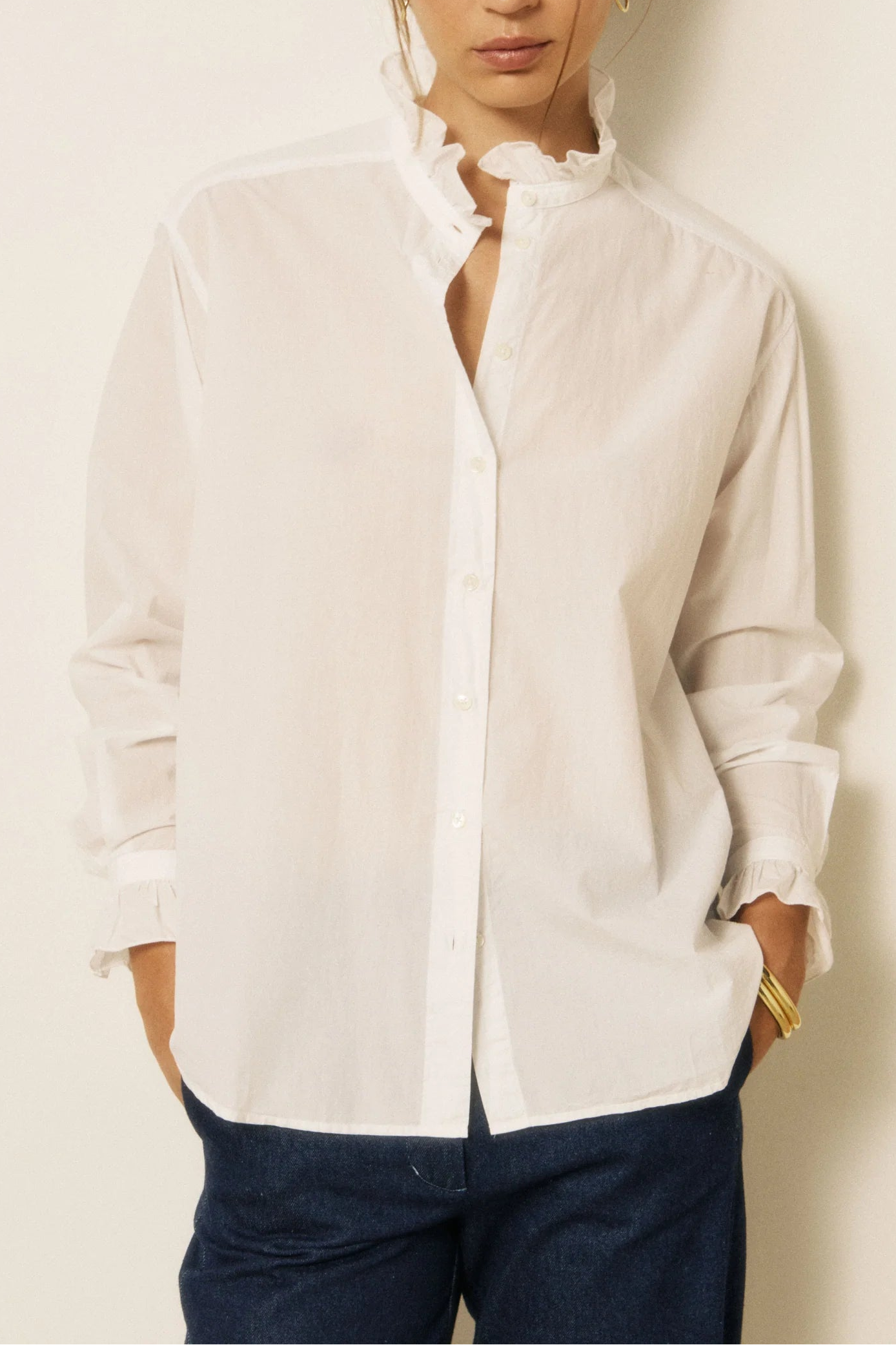 A woman modeling a white long-sleeve button-up shirt with a ruffled high collar, paired with dark blue jeans, against a plain off-white background.