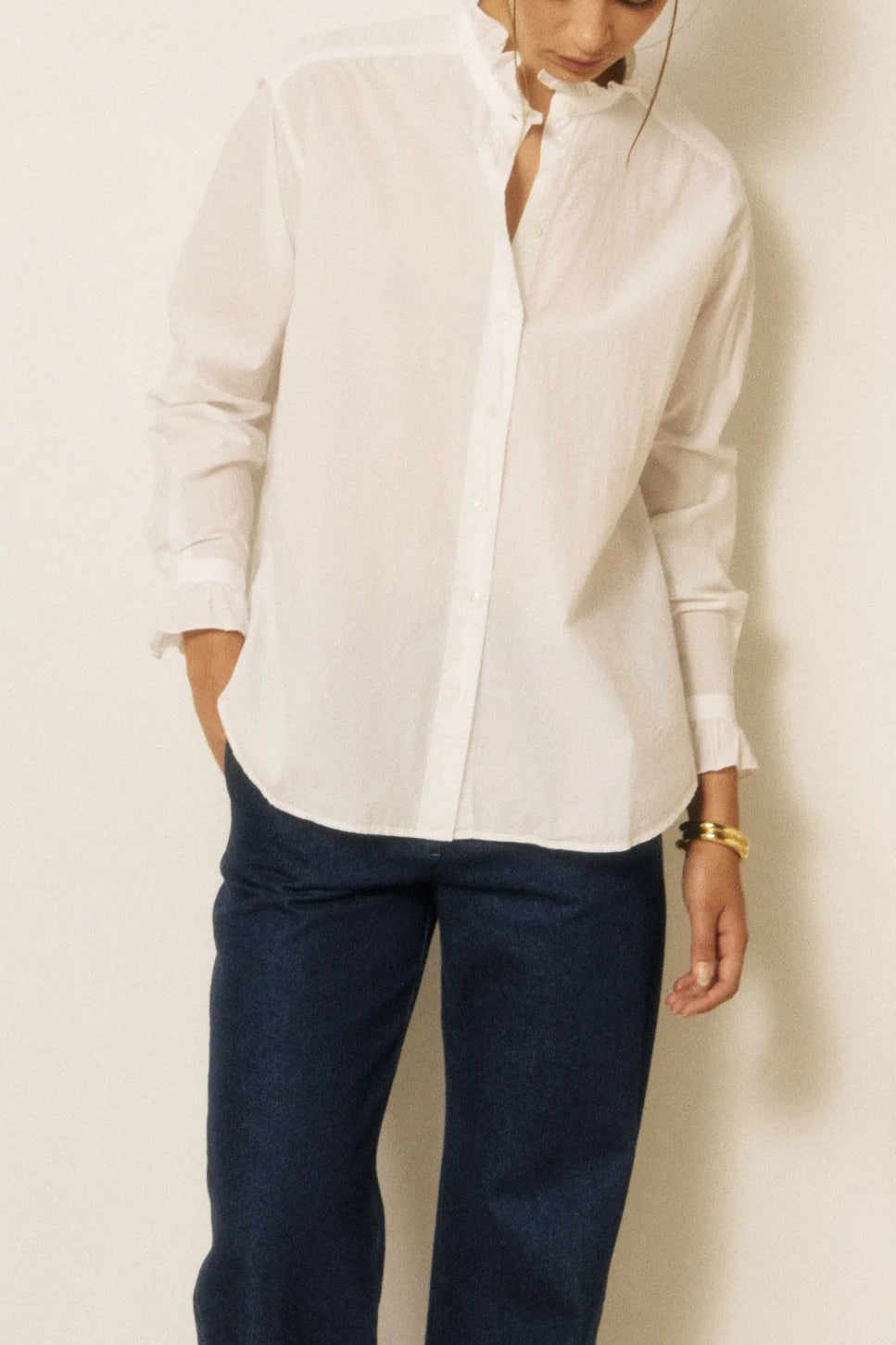 A woman modeling a white long-sleeve button-up shirt and dark blue wide-leg jeans, standing against a minimalist studio backdrop with a vertical wood panel.