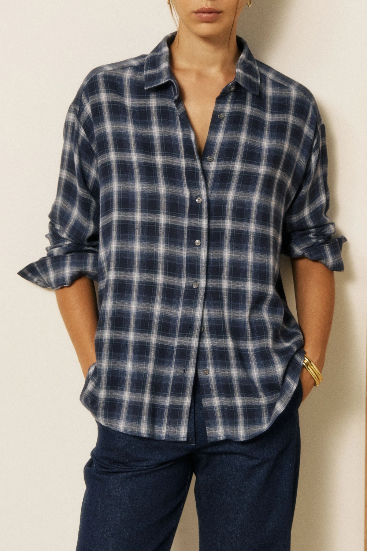 A woman wearing a navy and blue plaid button-down shirt with dark blue jeans, standing against a split cream and wood-paneled wall.