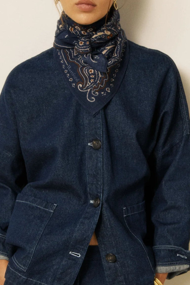 A model wearing a dark indigo denim jacket with a matching high-neck bandana scarf, posed against a neutral cream wall with a wooden panel on the right.