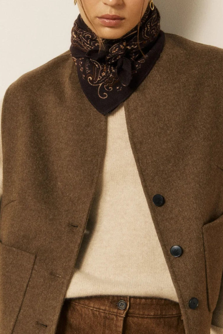 A model wearing a brown wool-blend vest over a cream knit sweater, paired with brown corduroy trousers and a dark patterned neck scarf, standing against a neutral off-white wall.