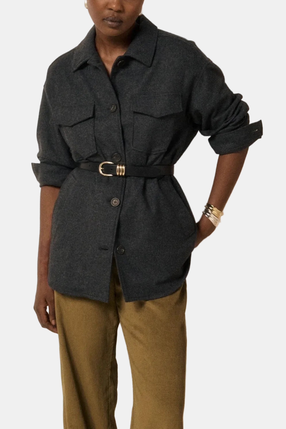 A model stands against a minimalist beige and wood-paneled wall, wearing a dark charcoal gray belted button-up shirt and olive green wide-leg trousers.