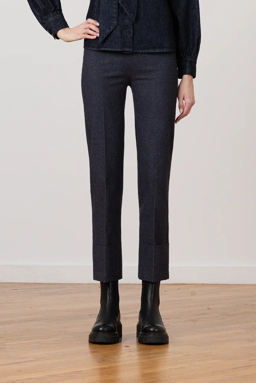 A woman stands in a studio wearing a dark navy textured jacket with a high collar and tie detail, matching dark navy trousers, and black ankle boots against a light gray wall and wood floor.