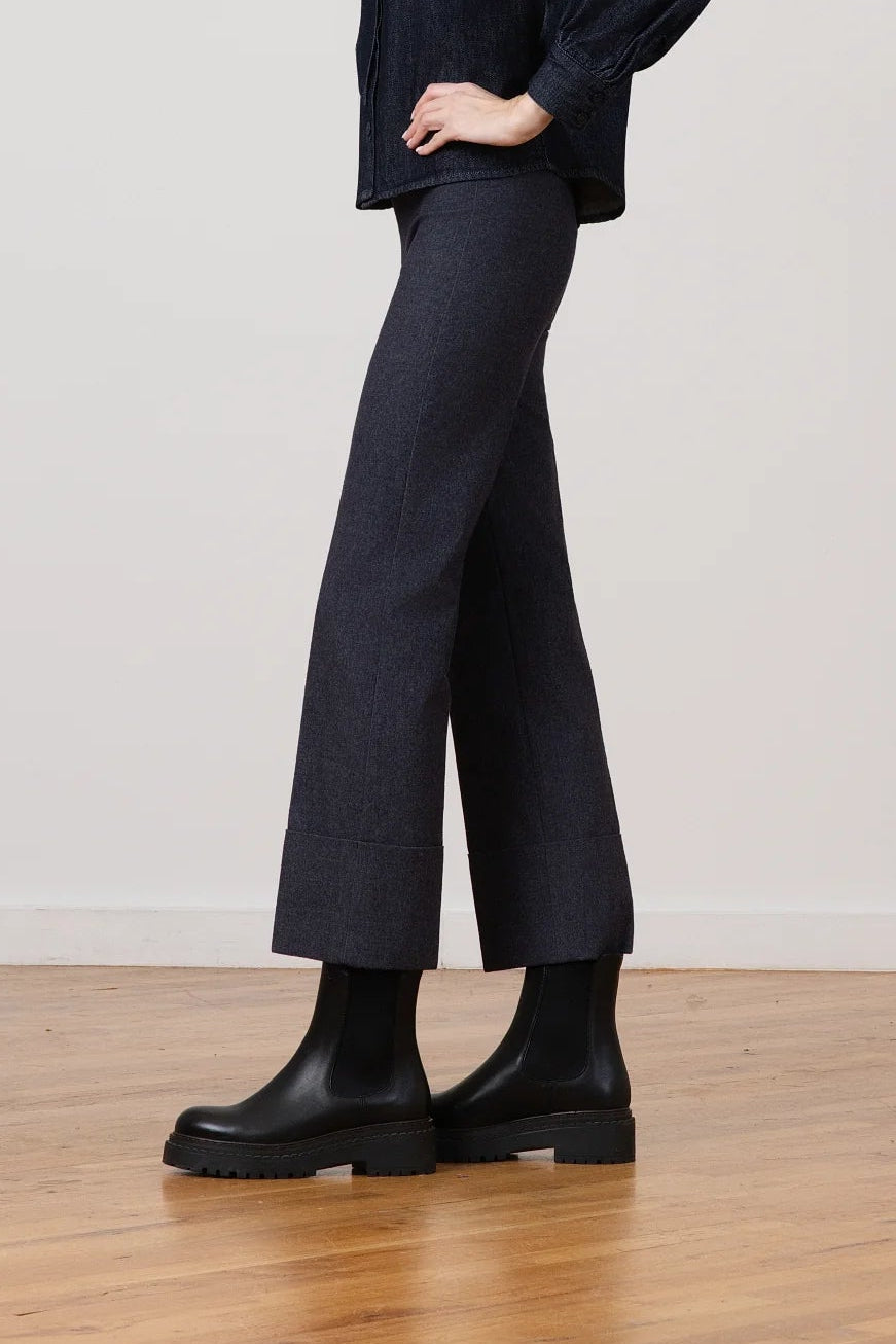 A woman modeling a dark navy textured jacket and matching wide-leg trousers with black boots, standing in a studio with a light gray wall and wooden floor.