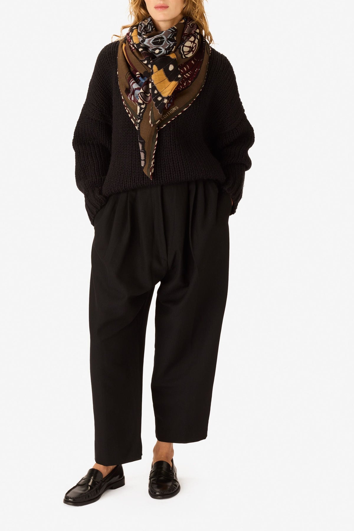 A model stands against a plain white studio background wearing a black knit sweater, black wide-leg trousers, black loafers, and a patterned scarf with brown, yellow, and purple tones.