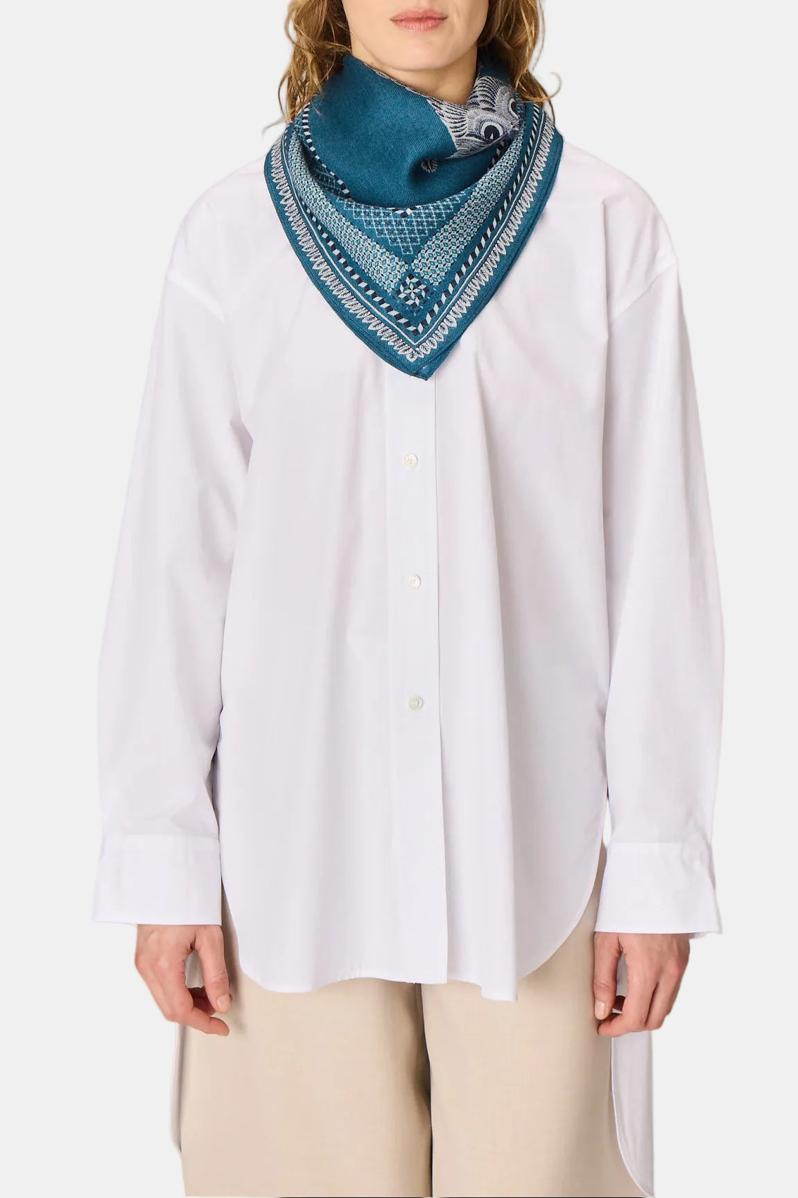 A woman wearing a white button-up shirt and beige pants, styled with a blue patterned bandana scarf, standing against a neutral light gray background.