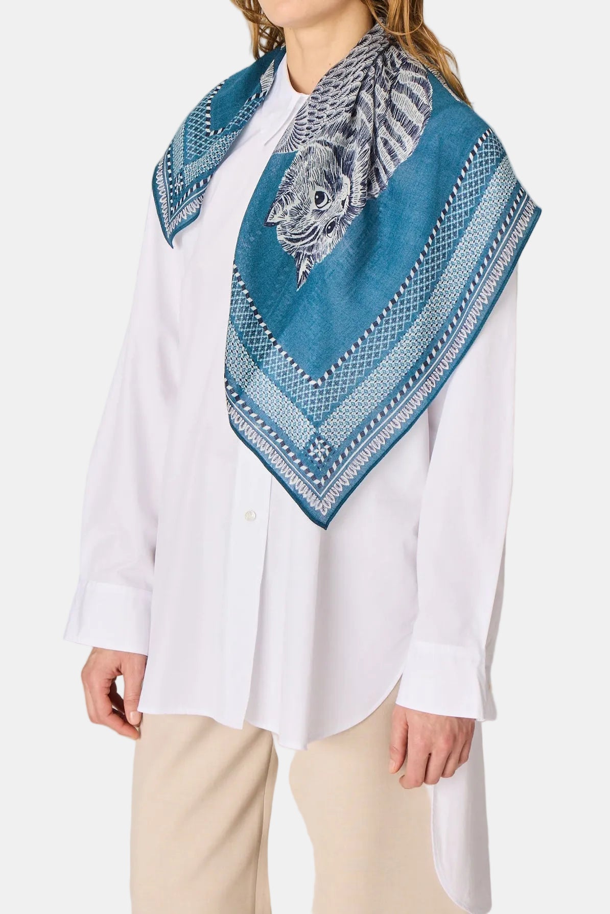 A woman wearing a white button-up shirt and beige trousers models a teal blue patterned square scarf with a black and white tiger design on a light gray studio background.