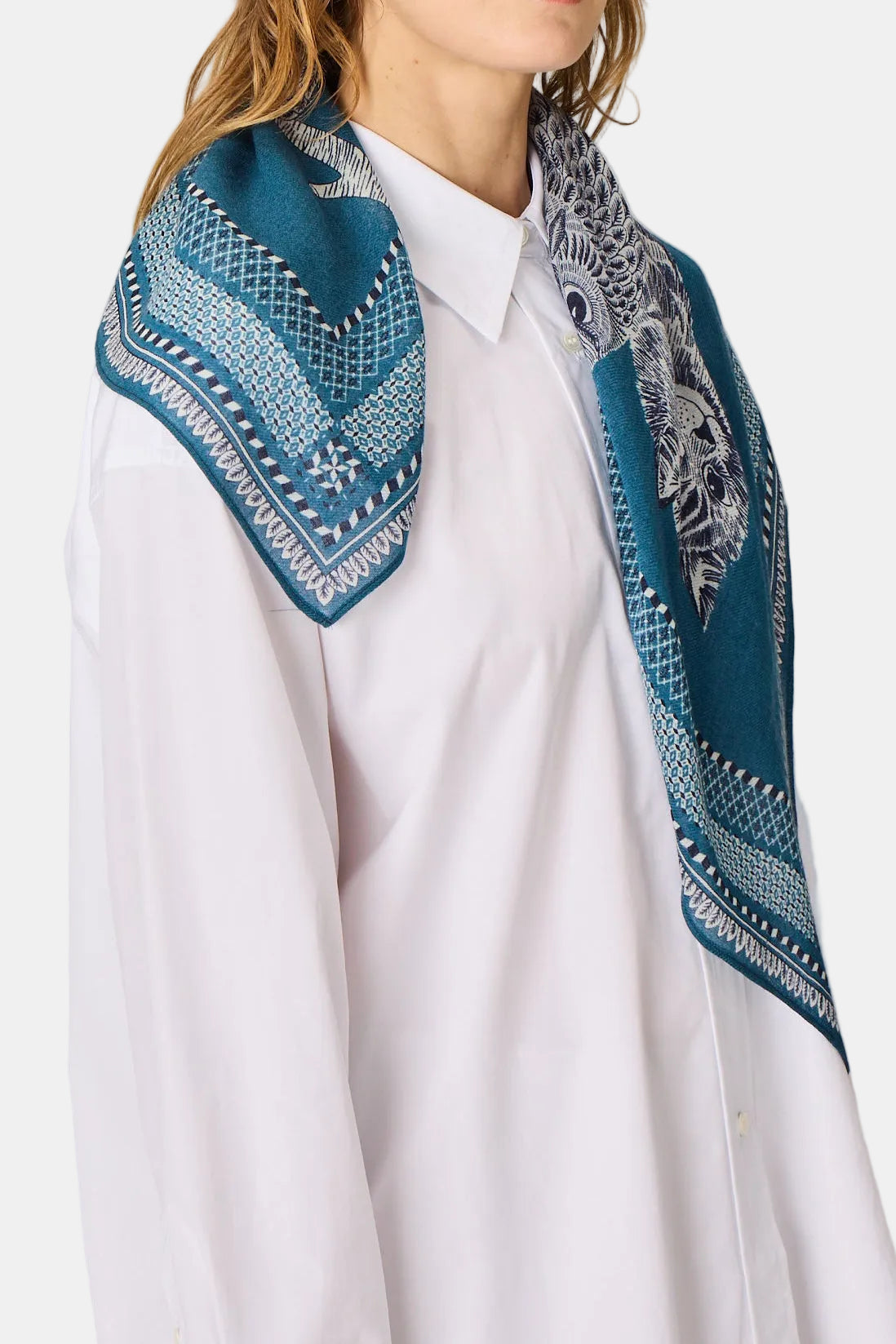 A woman with light brown hair wearing a white collared shirt and a teal blue patterned scarf, photographed against a solid light gray background.
