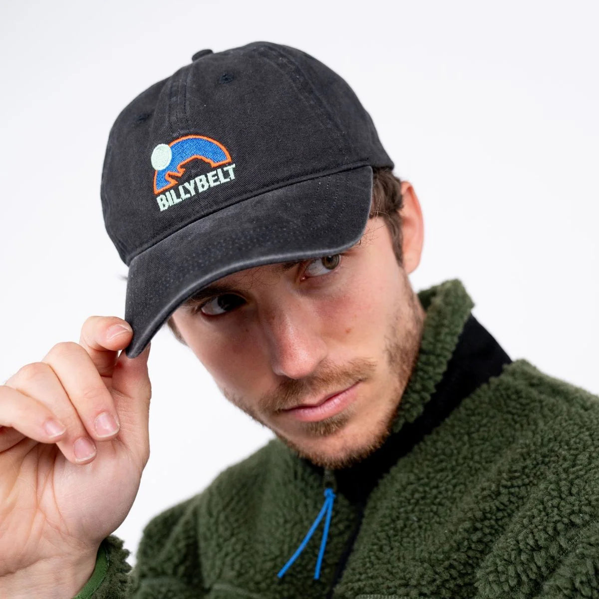 A man wearing a black washed baseball cap with a blue and orange embroidered mountain logo and the text 'BILLYBELT' on the front, holding the brim with one hand while wearing an olive green fleece jacket against a plain white background.