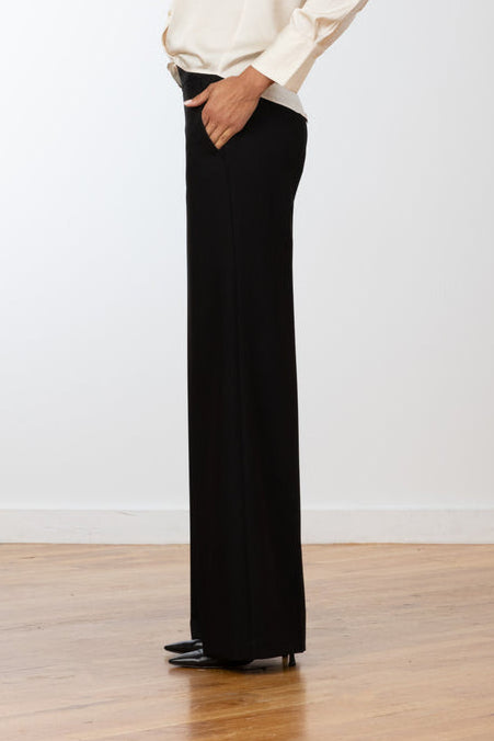 A woman stands in profile wearing a cream long-sleeve blouse and black wide-leg pants against a plain white wall and light wood floor.