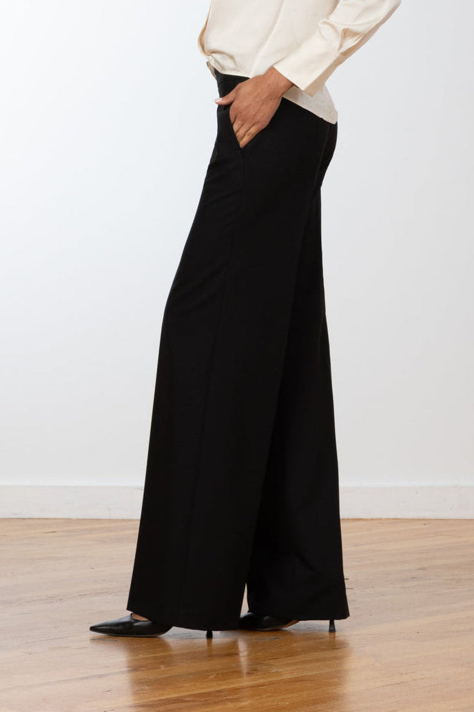 A woman stands in profile wearing a cream long-sleeve blouse and black wide-leg trousers, posed on a light wood floor against a plain white wall.