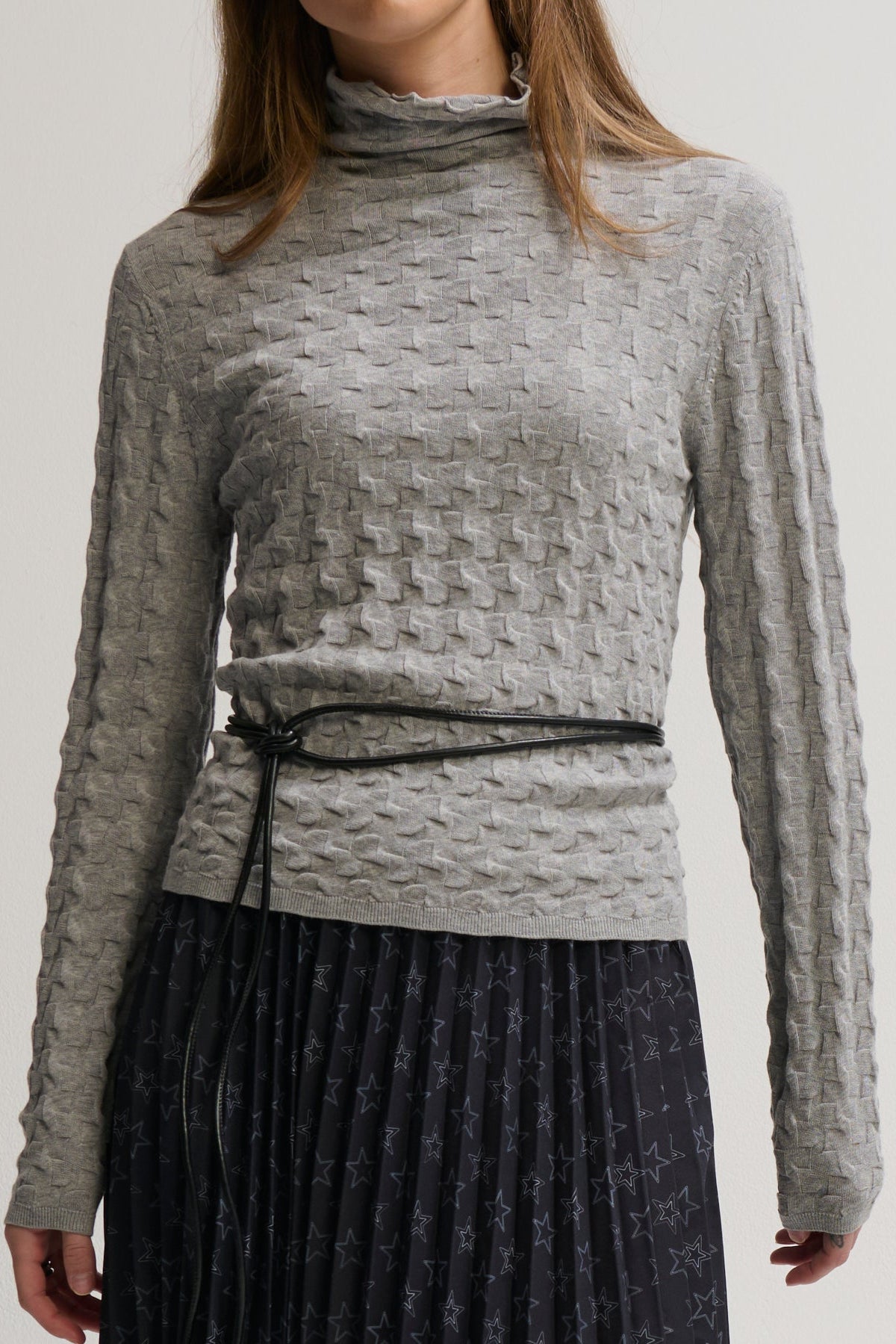 Person wearing a textured gray long-sleeve top with a belt, standing against a plain background.