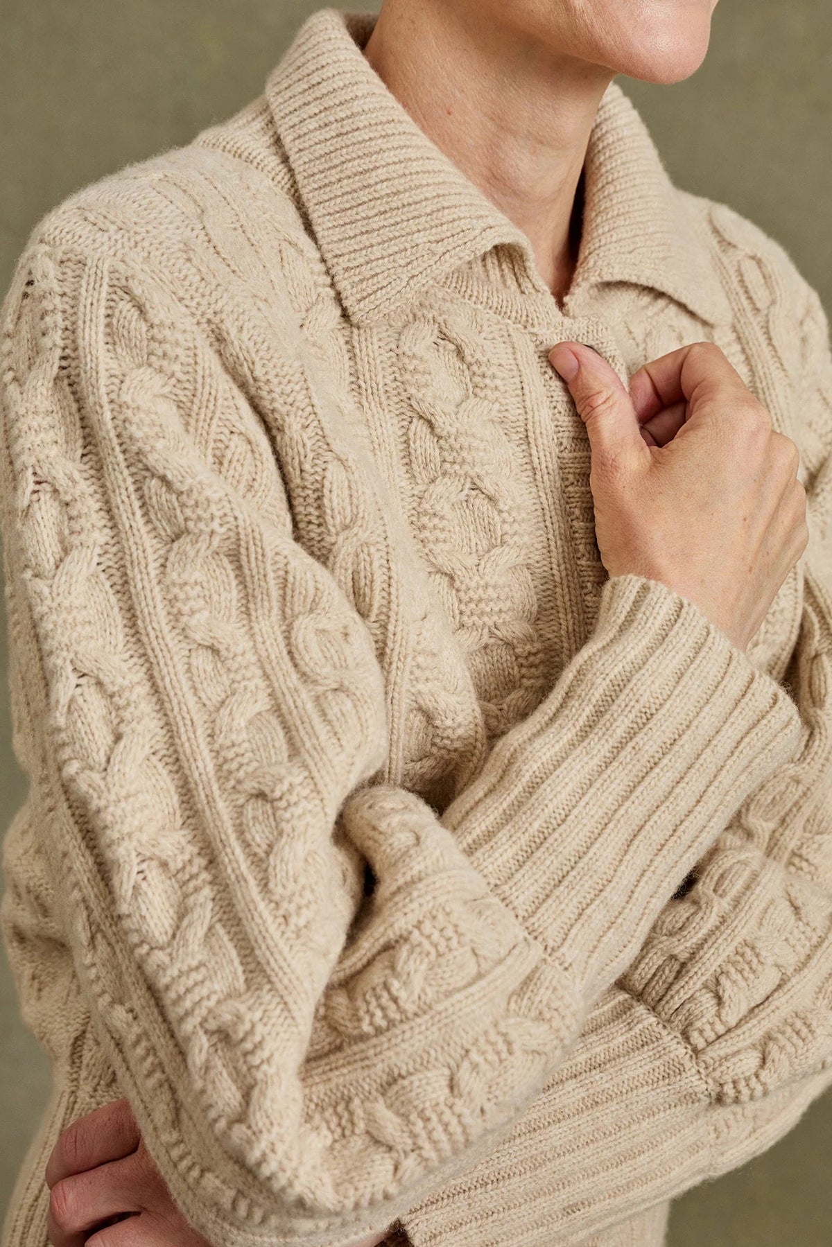 A close-up of a person wearing a chunky cable-knit sweater in a warm cream or oatmeal beige color, with a collar and ribbed cuffs and hem.