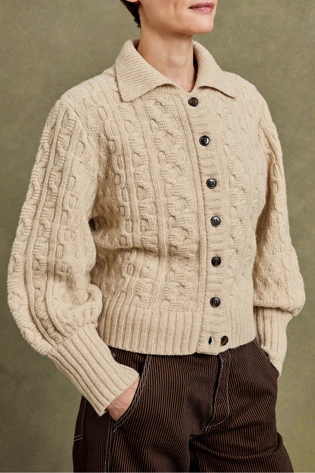 A model with short dark hair wearing a cream-colored cable-knit button-up cardigan and brown corduroy trousers, standing against a textured olive-green studio background.