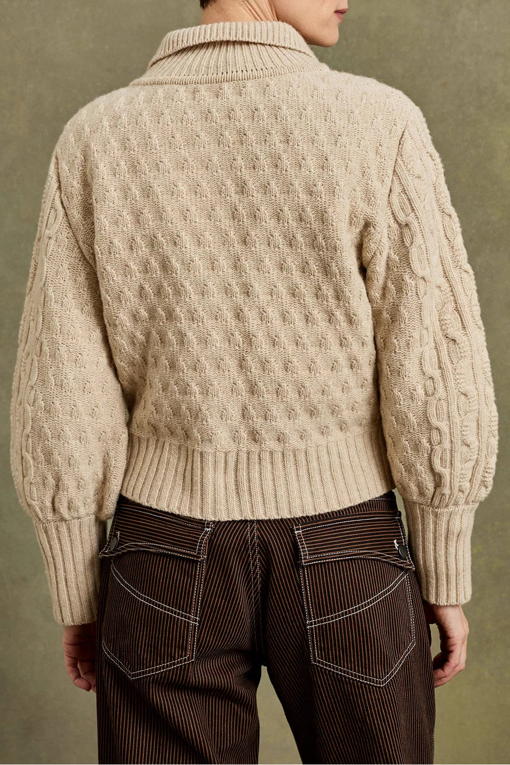 A model shown from the back wearing a cream-colored cable-knit sweater and dark brown corduroy trousers, set against a textured olive-green studio background.