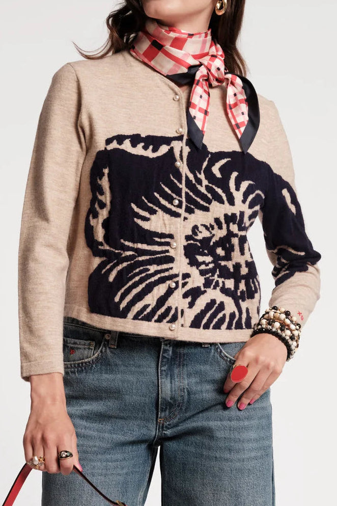 A woman models a beige and black patterned knit sweater, paired with blue jeans, a red and white scarf, and holding a red leather shoulder bag against a plain white background.