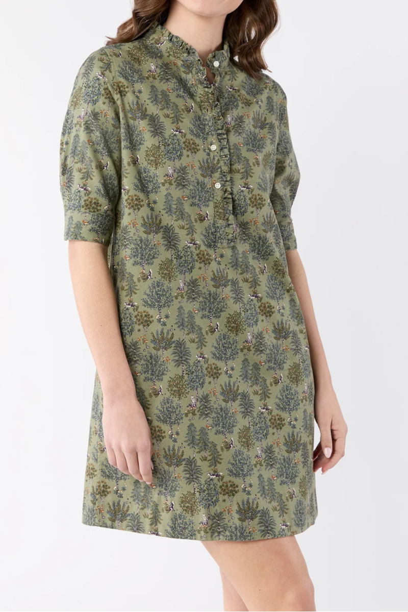 A woman modeling a short-sleeved button-front dress with an allover botanical print in olive green, beige, and muted blue-gray, photographed against a plain light gray studio background.