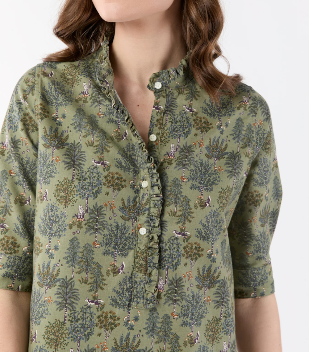 A woman wearing a short-sleeved button-up shirt with a detailed botanical and bird pattern in olive green, brown, and beige, photographed against a plain white background.