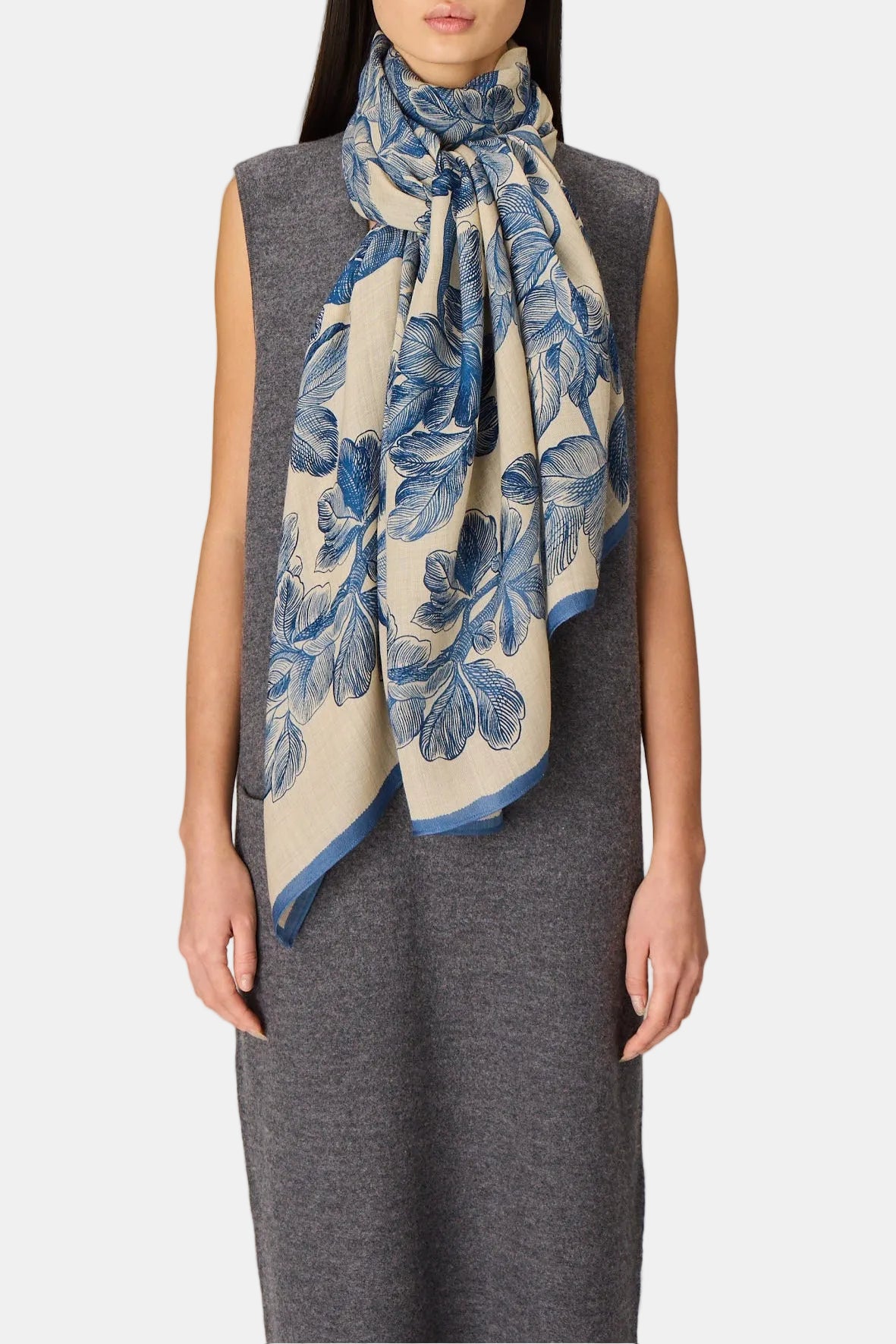 A model wearing a gray sleeveless dress and a cream-colored scarf with a blue floral pattern, standing against a neutral light gray background.