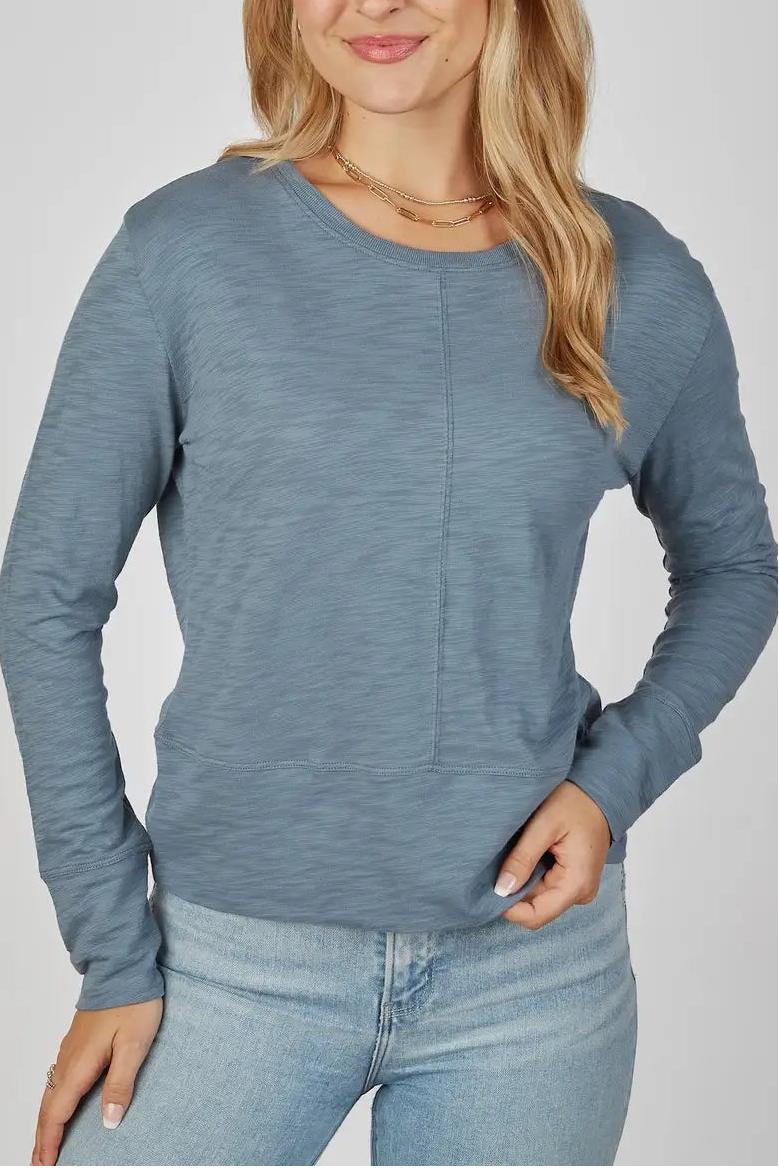 A woman modeling a slate blue long-sleeve crewneck top with a subtle vertical seam detail on the chest, paired with light-wash blue jeans, against a solid light gray background.
